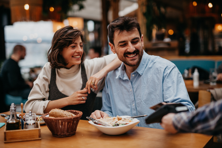 Spontaneous image of a beautiful brunet enjoying lunch with her friend, a birded guy, both casually dressed. A man is paying with a card the lunch, both smiling